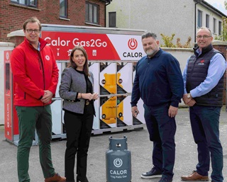 Four individuals standing in front of a Calor Gas2Go vending station, which features multiple compartments storing yellow LPG cylinders. A Calor 11kg Patio Gas cylinder is placed on the ground in front of them. The outdoor setting near residential buildings suggests a promotional event for Calor’s automated gas cylinder service.