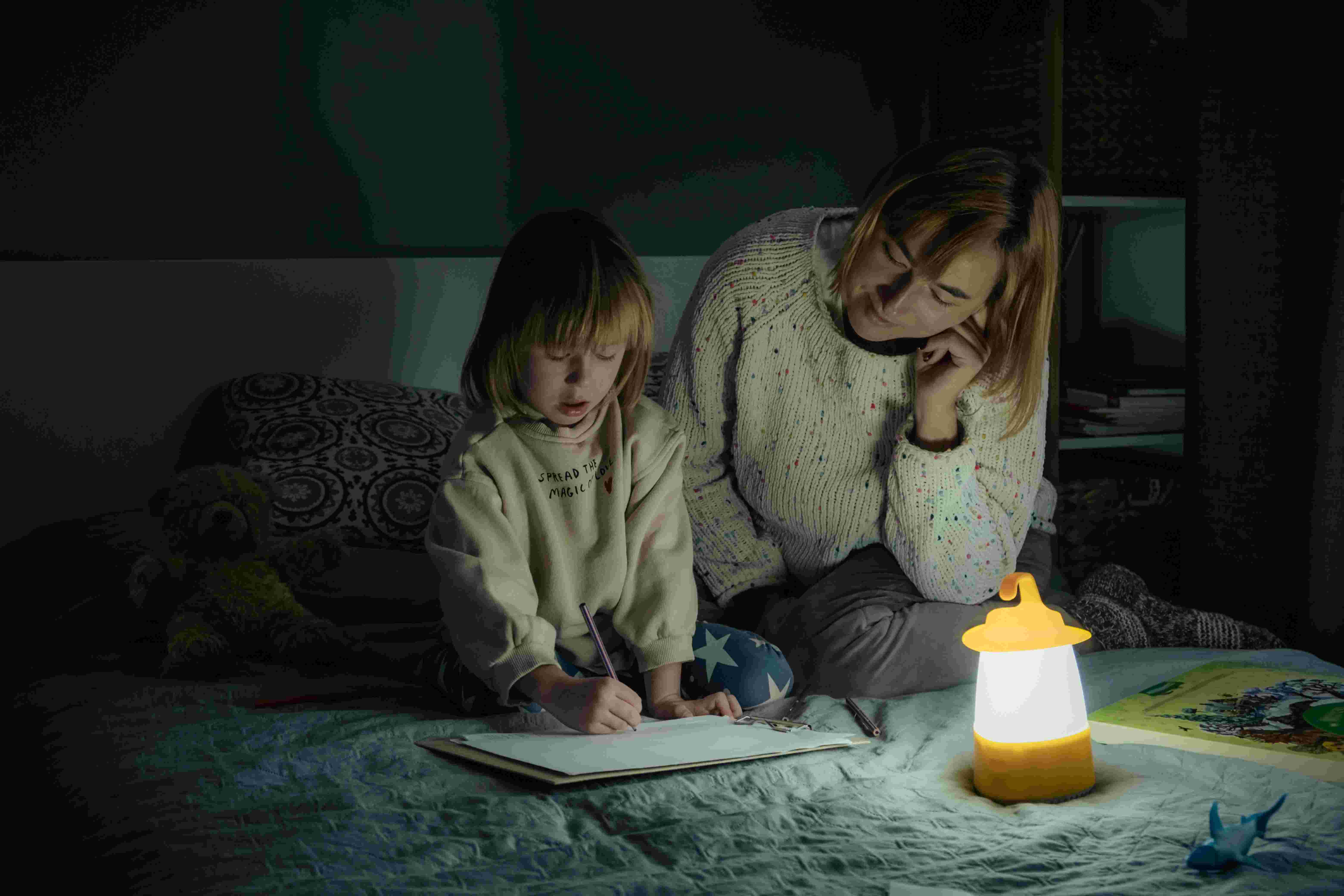 Mother and little daughter studying and drawing in a complete darkness during electricity outage. Little girl uses camping lantern to do her homework during blackout. Energy crisis concept