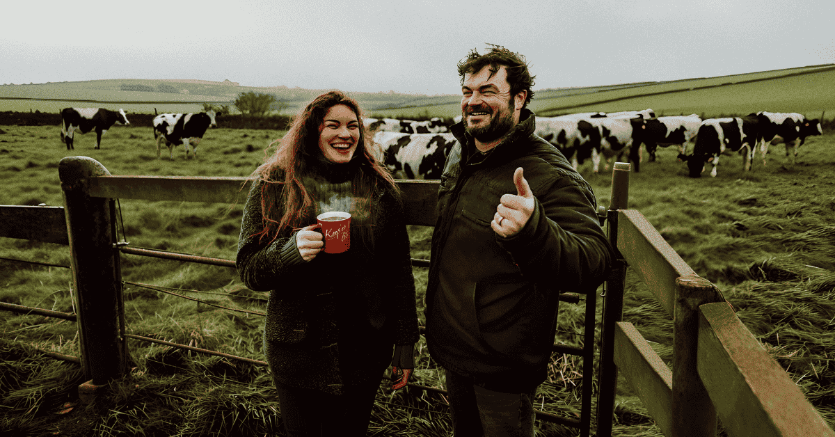 A man and woman standing in front of a green field with cows, the couple are smiling holding a cup of tea