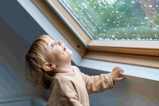 Little preschool boy staying home in bad weather and looking with interest through window on raindrops and hail, idoors.